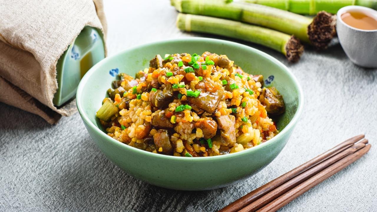 A ceramic bowl filled with pork and bamboo shoot fried rice, placed on a wooden kitchen counter. The rice is glossy with soy sauce, topped with a sprinkle of green onions. Next to the bowl are wooden chopsticks and a small cup of green tea. In the background: a half-used bag of jasmine rice, a jar of oyster sauce, and a bunch of fresh bamboo shoots. Natural light streams through a nearby window, giving the scene a soft, inviting glow.