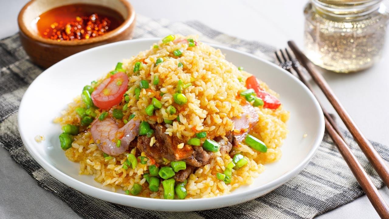 Inviting finished fried rice: Fluffy golden rice mounded on a white ceramic plate, topped with char siu, pink shrimp, bright green peas, and green onions. A small ramekin of sriracha sits beside it. Background shows a family dining table with a checkered cloth, wooden chopsticks, and a mason jar of iced tea. Natural light makes the rice glow, highlighting the vibrant colors of the ingredients. The scene feels warm, like a Sunday brunch with loved ones.